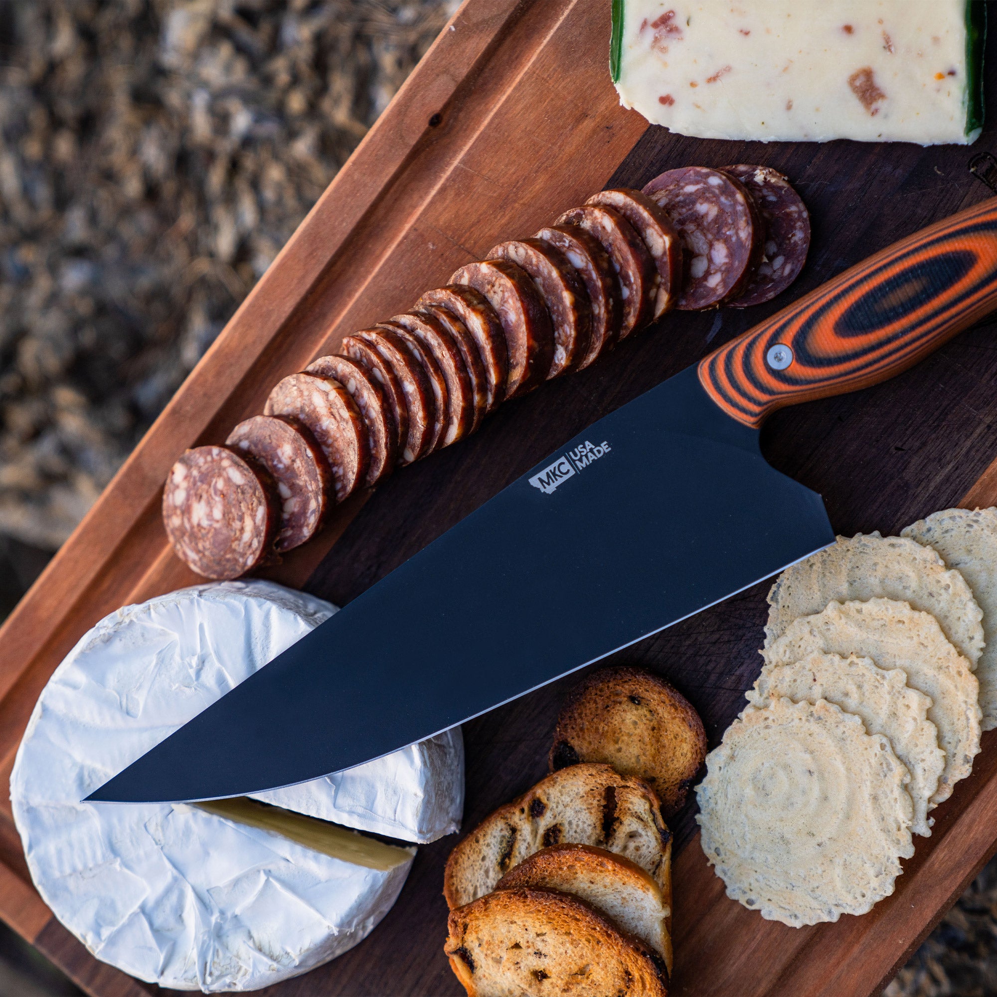 Alt text: "Bighorn Chef orange and black handled knife on a wooden board, displayed with sliced sausage, cheese, and toasted bread, outdoors."