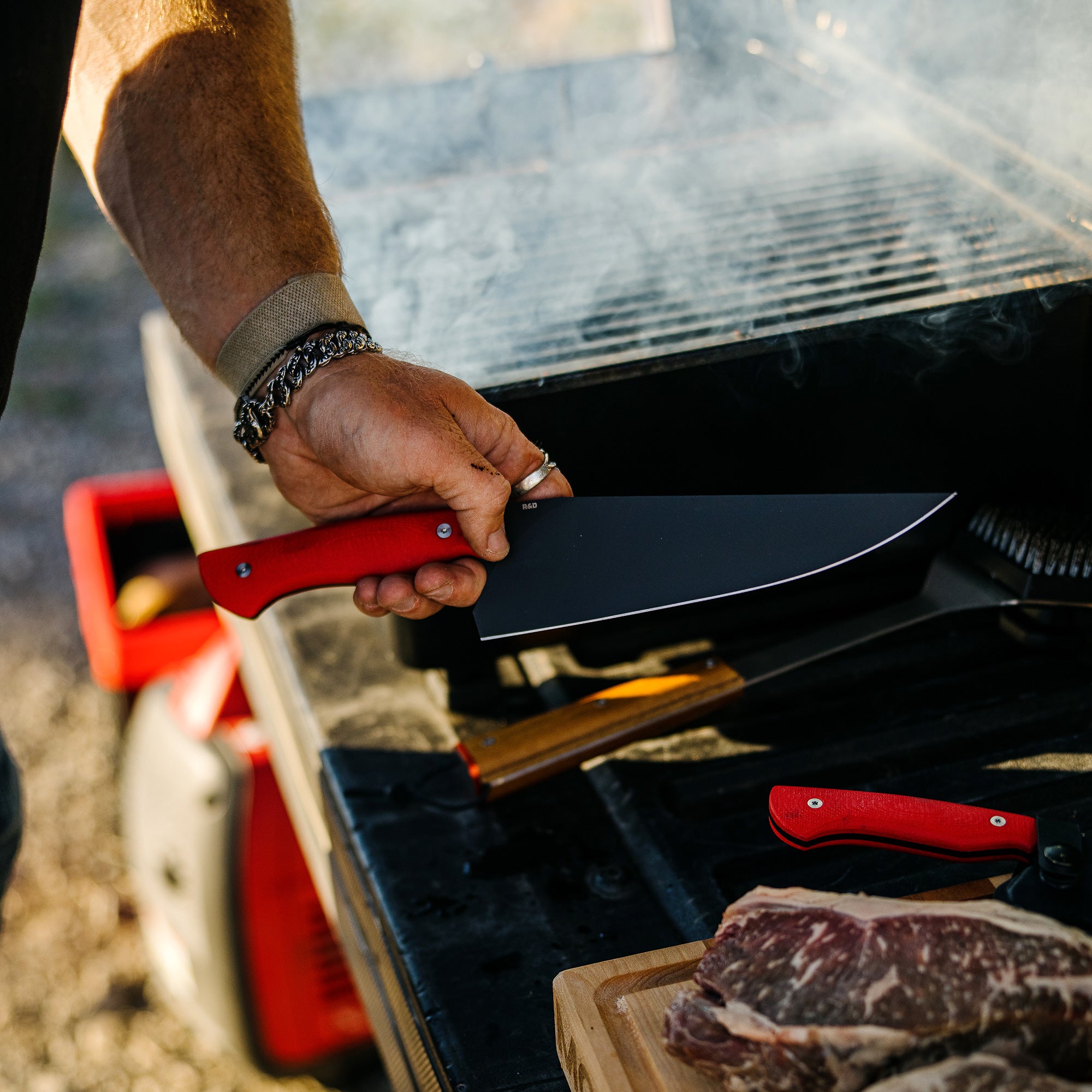 Close-up of a hand holding a large Bighorn Chef knife with a red handle over a grill, with other red-handled knives on a wooden board next to a piece of raw steak, outdoors.