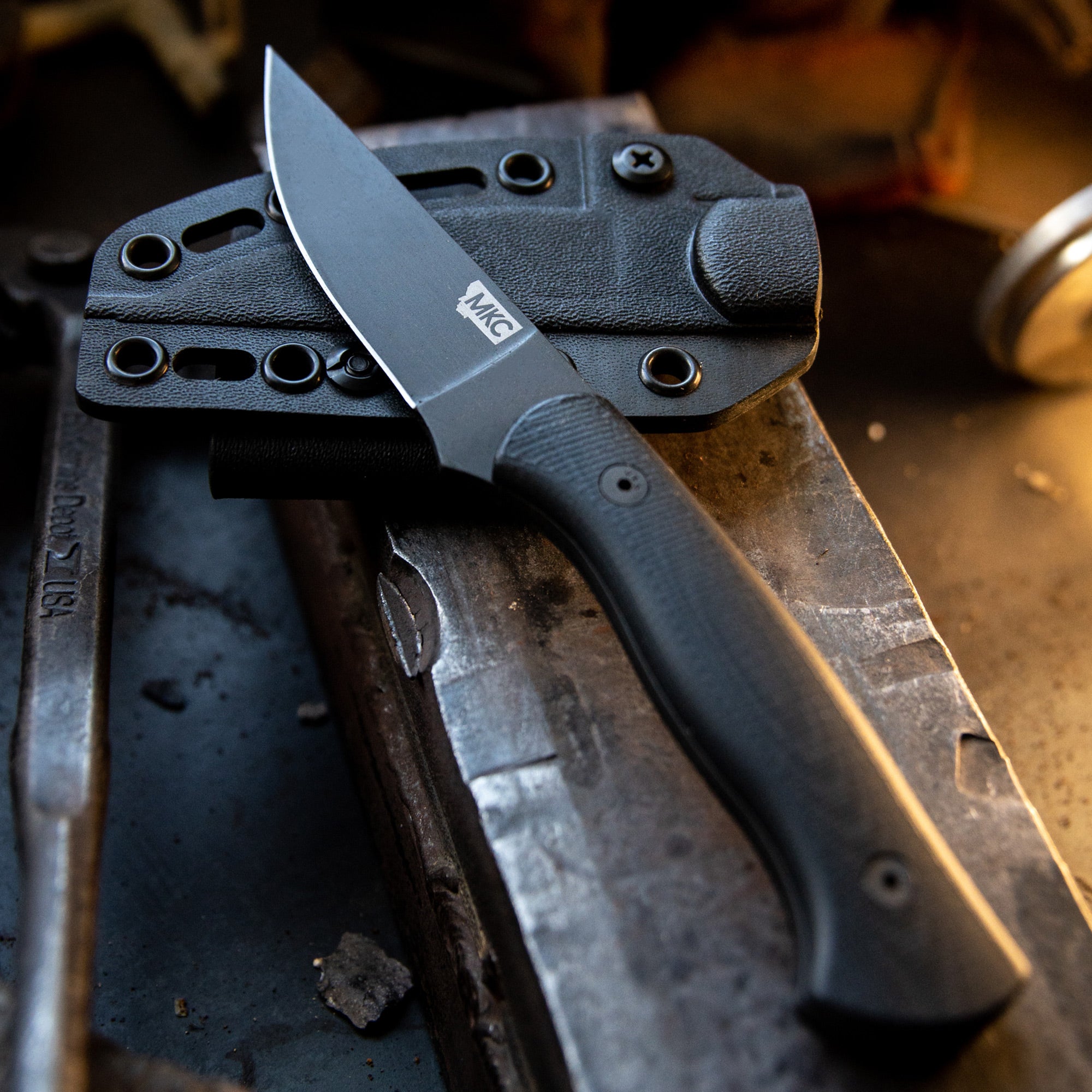 Alt text: "Durable MKG Blackfoot 2.0 tactical knife with a black blade and handle, displayed on a textured, metallic workshop surface alongside its custom black sheath with multiple attachment points."