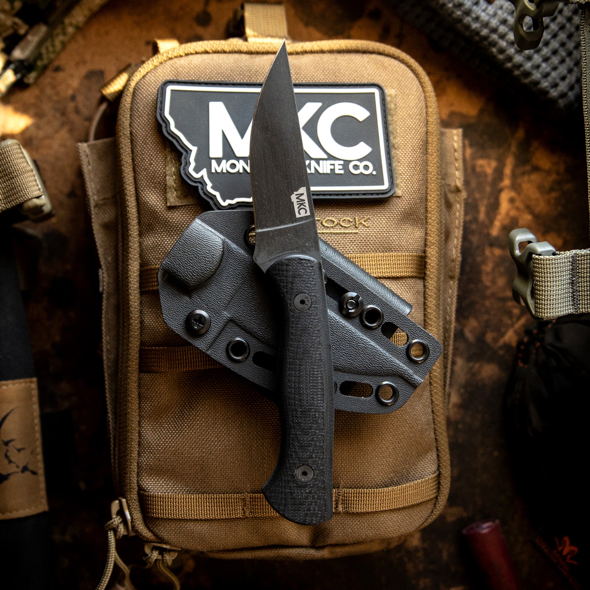 Alt Text: "BLACKFOOT 2.0 tactical knife by MONOCK Knife Co. displayed with a black handle and sheath, attached to a khaki tactical backpack with the company logo visible."