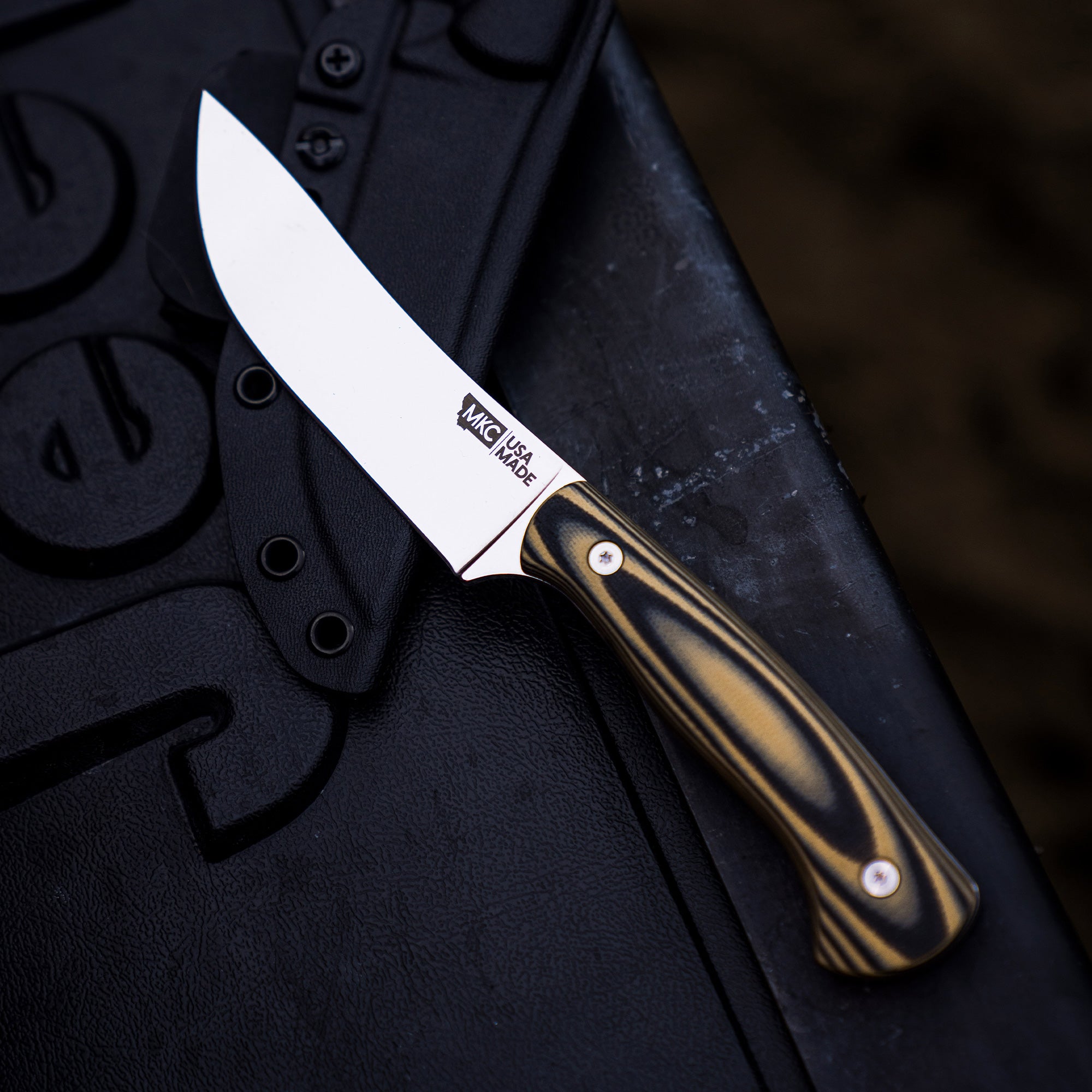 Alt text: A MagnaCut Stonewall Skinner knife by the Mule Deer Foundation, featuring a sleek blade with "MKC USA MADE" engraved, and a polished handle with striped patterns, displayed on a textured black surface.