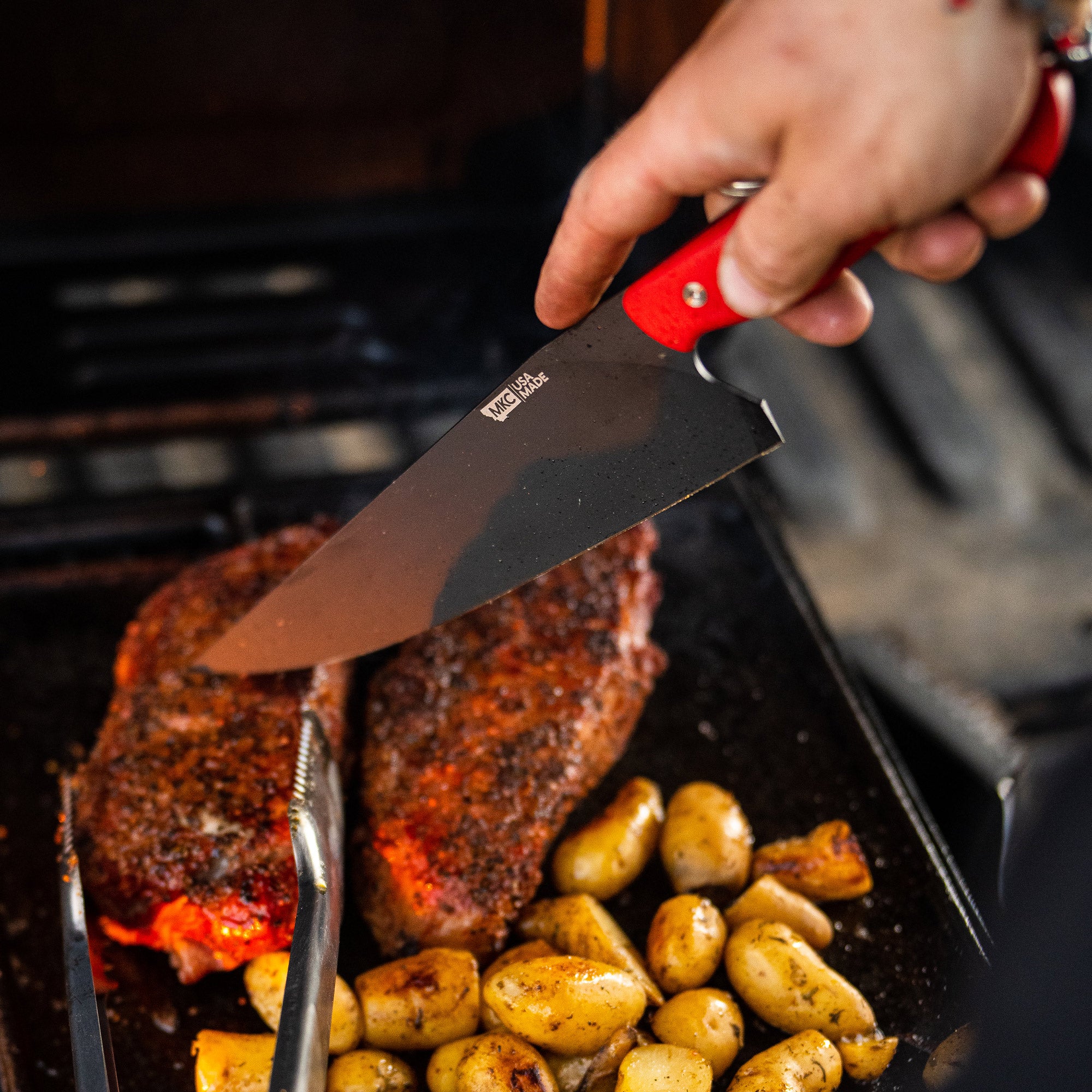 Alt text: "Close-up of a red-handled BIGHORN CHEF knife slicing through a juicy steak in a pan, surrounded by roasted potatoes, emphasizing the sharpness and quality of the knife."