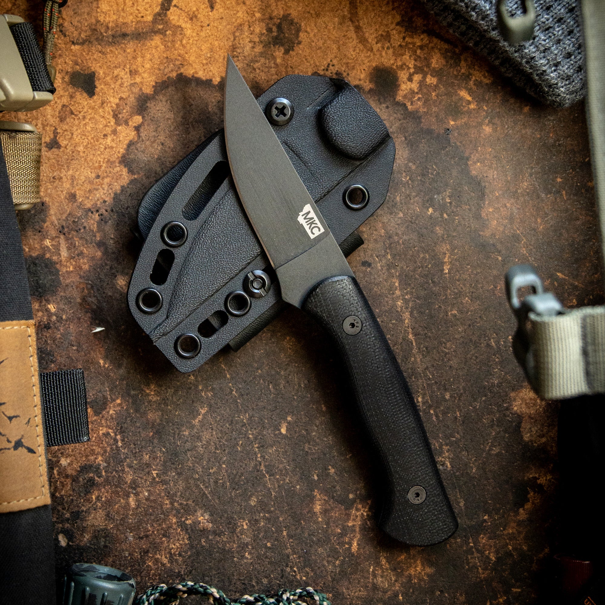 Alt text: "Blackfoot 2.0 tactical knife in black with a textured handle and sheath, displayed on a rustic metal background surrounded by tactical gear."