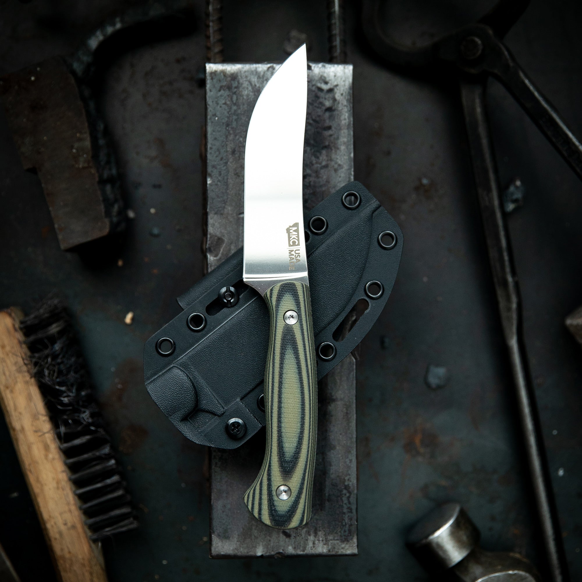 High-quality image of a limited-edition Mule Deer Foundation MagnaCut Stonewall Skinner knife displayed with its black Kydex sheath, featuring a Micarta handle with green and black layers, set against a rustic, industrial background with tools.