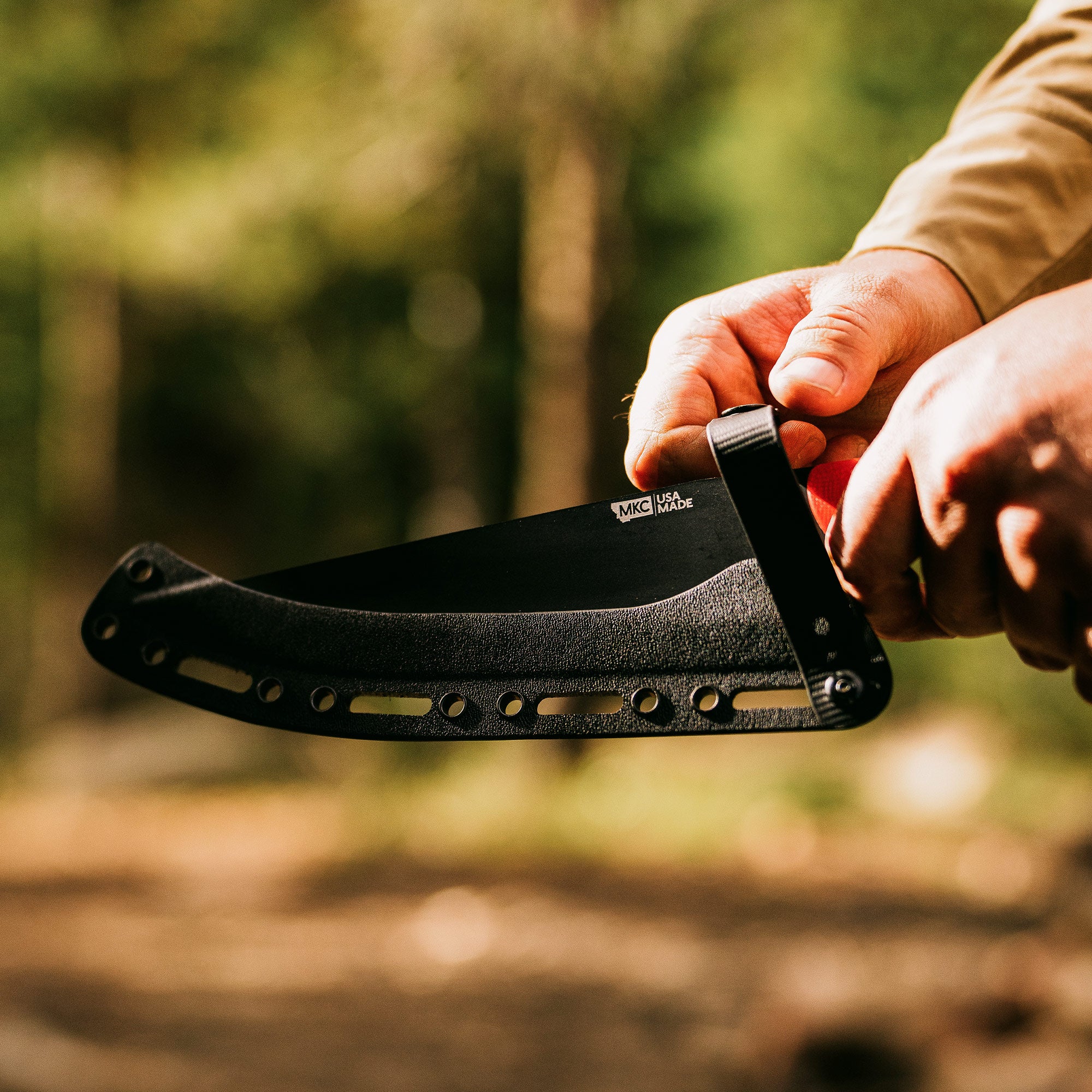 Alt text: "Close-up of a person's hand holding the Bighorn Chef red handle knife with a black blade, featuring multiple rivets and stamped with 'MKC USA MADE', set against a blurred forest background."