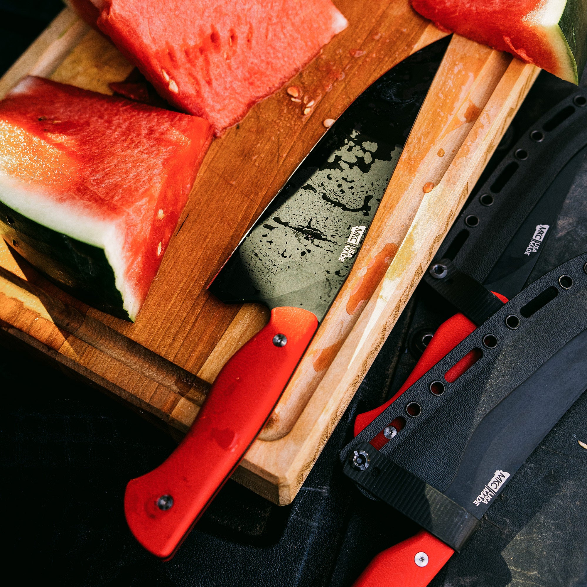 Alt text: "A Bighorn Chef red-handled knife alongside various other knives, displayed on a wooden cutting board with freshly cut watermelon slices, emphasizing professional kitchen tools."