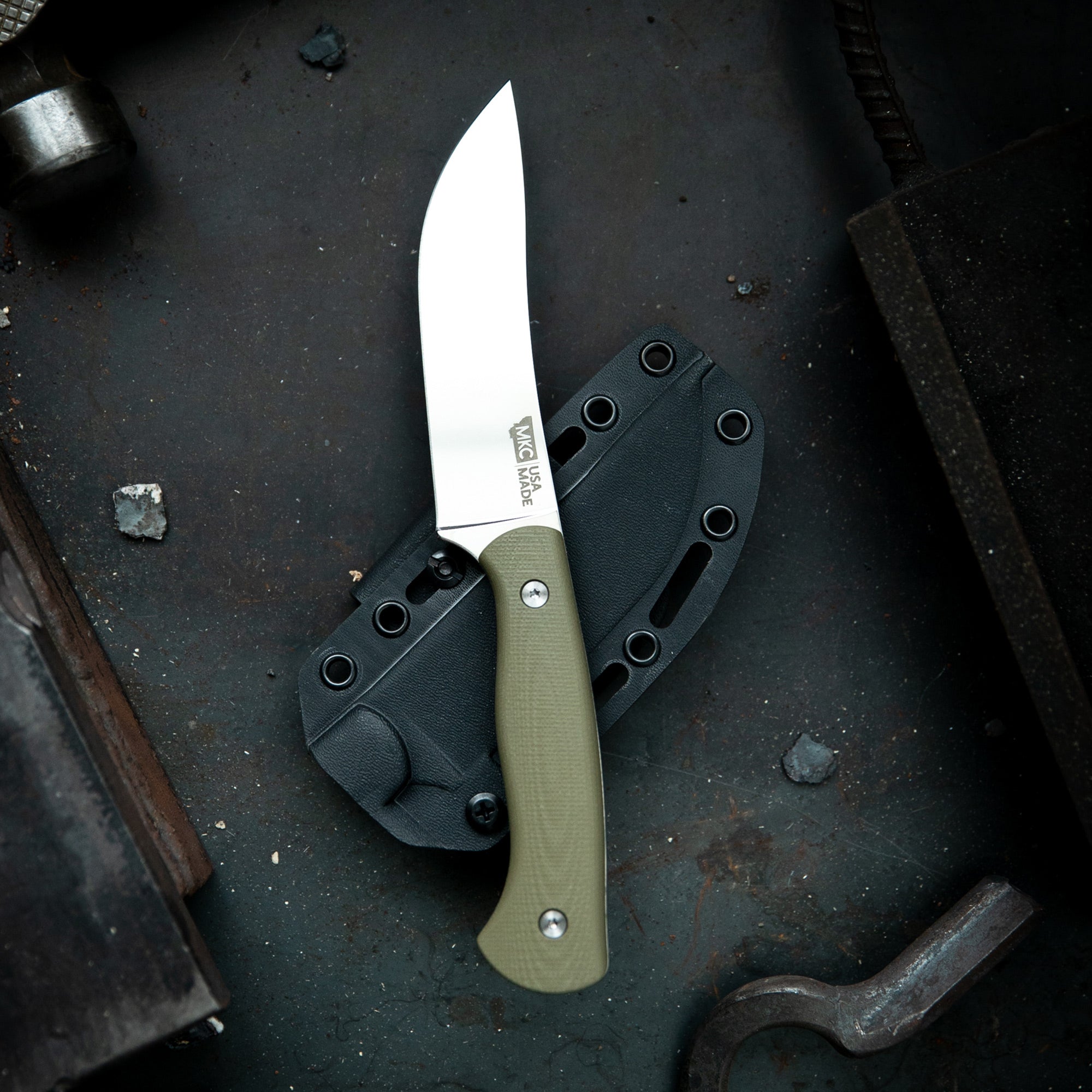 Alt text: Mule Deer Foundation MagnaCut Stonewall Skinner knife with a sharp, straight blade and a textured olive green handle, displayed alongside its black sheath on a dark, gritty workshop background.