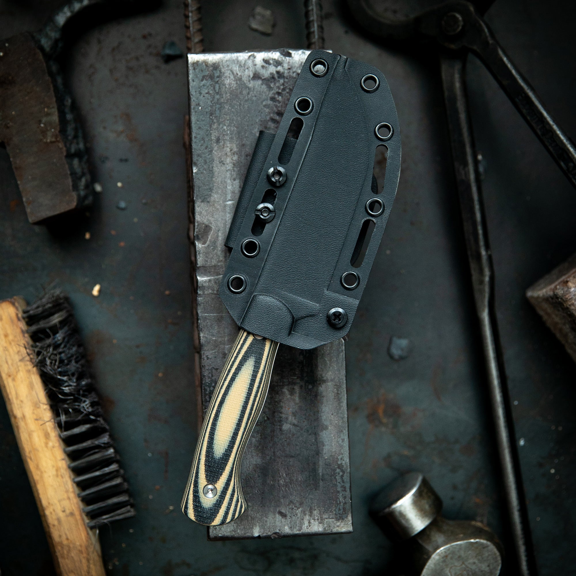 Alt text: "Magnacut Stonewall Skinner knife with a striped handle, housed in a black kydex sheath, displayed on a rustic metal workshop table surrounded by tools."