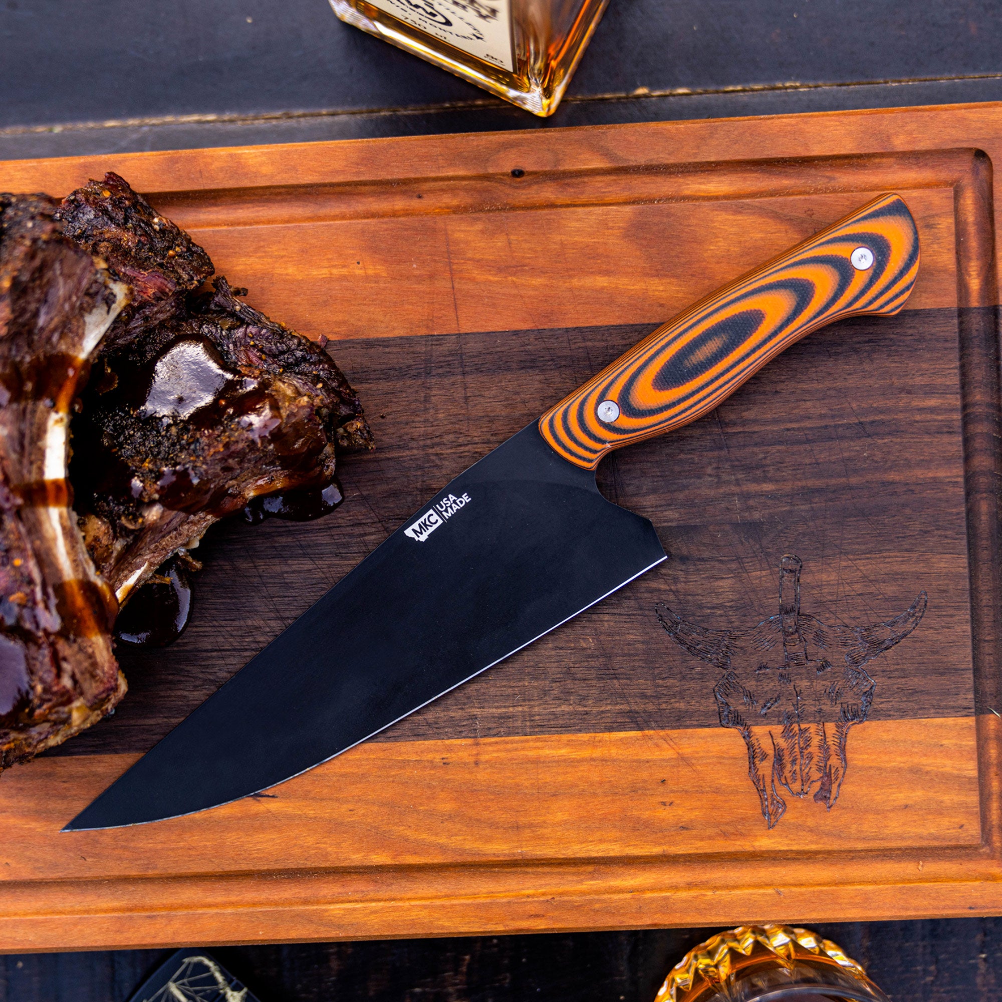Alt text: "High-quality chef's knife with a sleek black blade and vibrant orange and black handle, displayed on a wooden board alongside a cooked rack of ribs and a bottle. Ideal for culinary presentations."