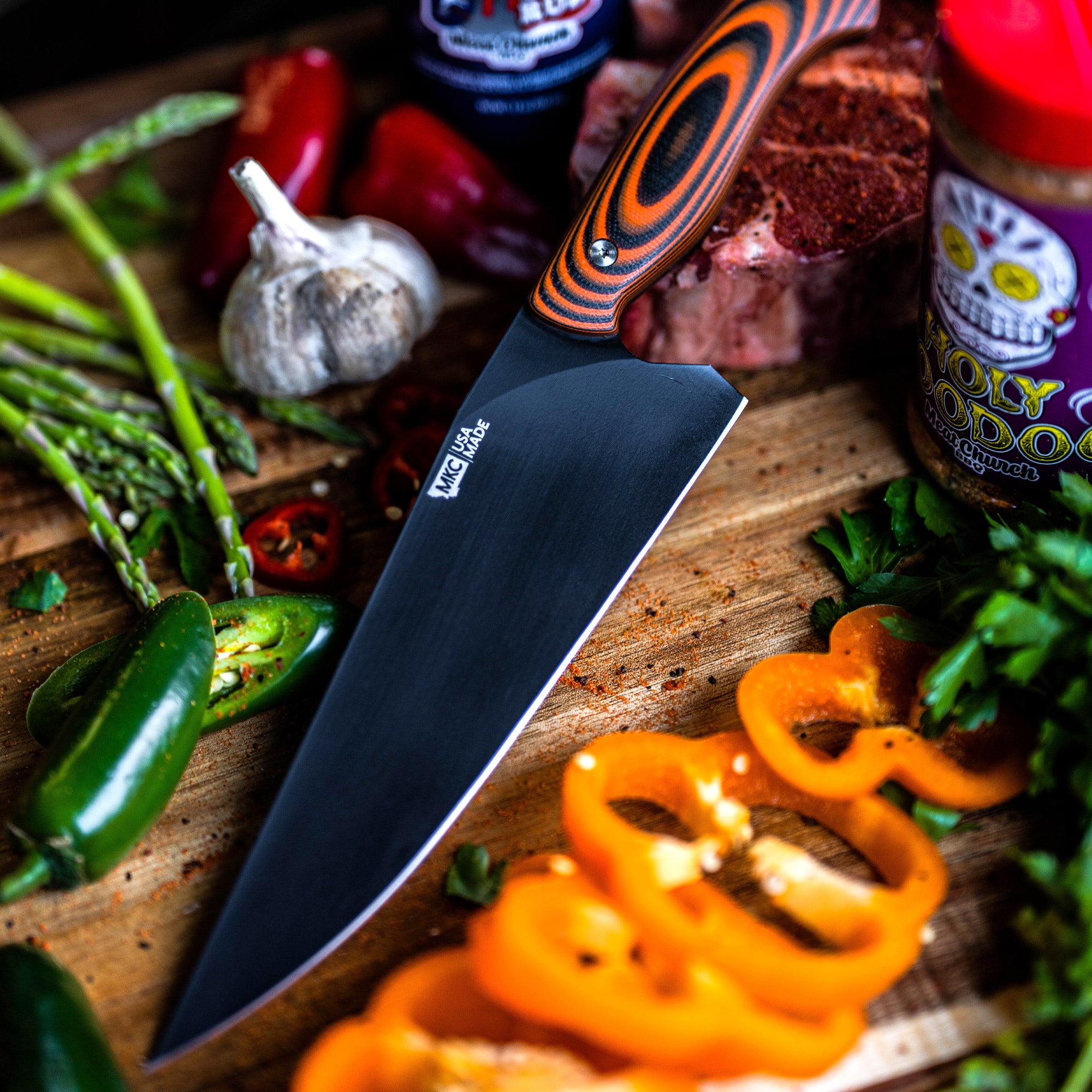 A Bighorn Chef kitchen knife with orange and black handle, displayed on a cutting board amidst an array of fresh vegetables including orange bell peppers, jalapeños, and asparagus.