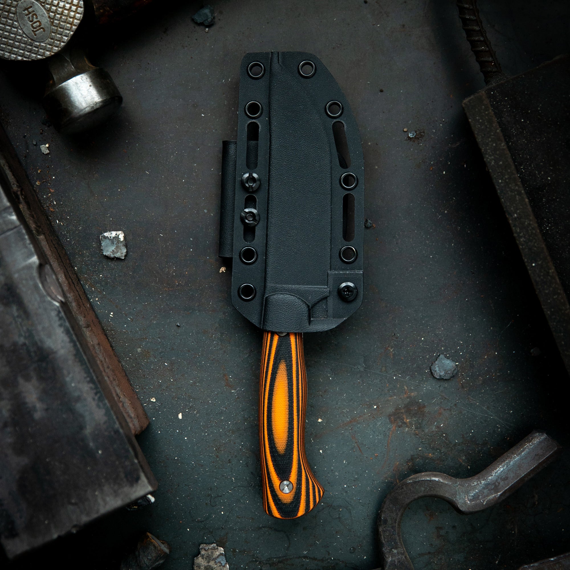 Alt text: Black Magnacut Stonewall Skinner knife in a sheath, featuring an orange-striped handle, displayed on a gritty workshop surface with vintage tools around. This is a limited edition item from the Mule Deer Foundation, labeled as sold