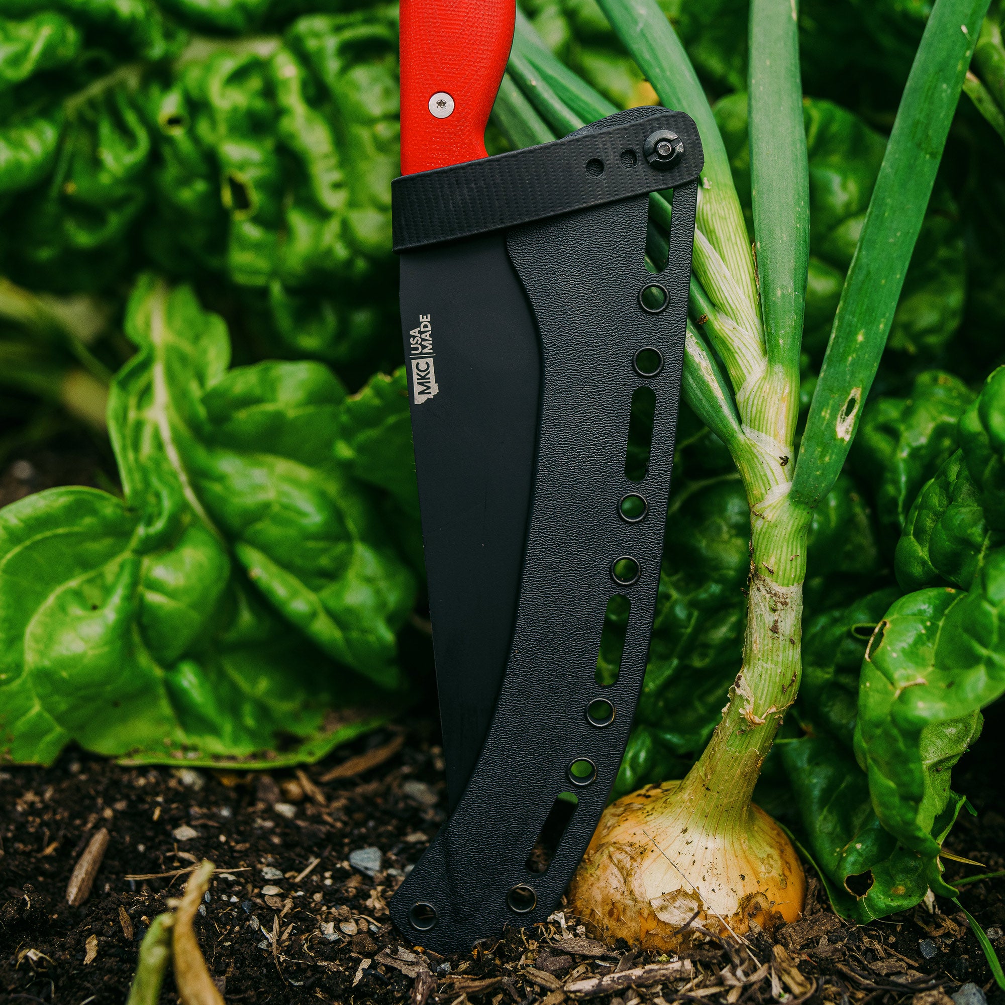 "BIGHORN CHEF red-handled knife with a black sheath, partially embedded in soil near fresh vegetables in a garden."