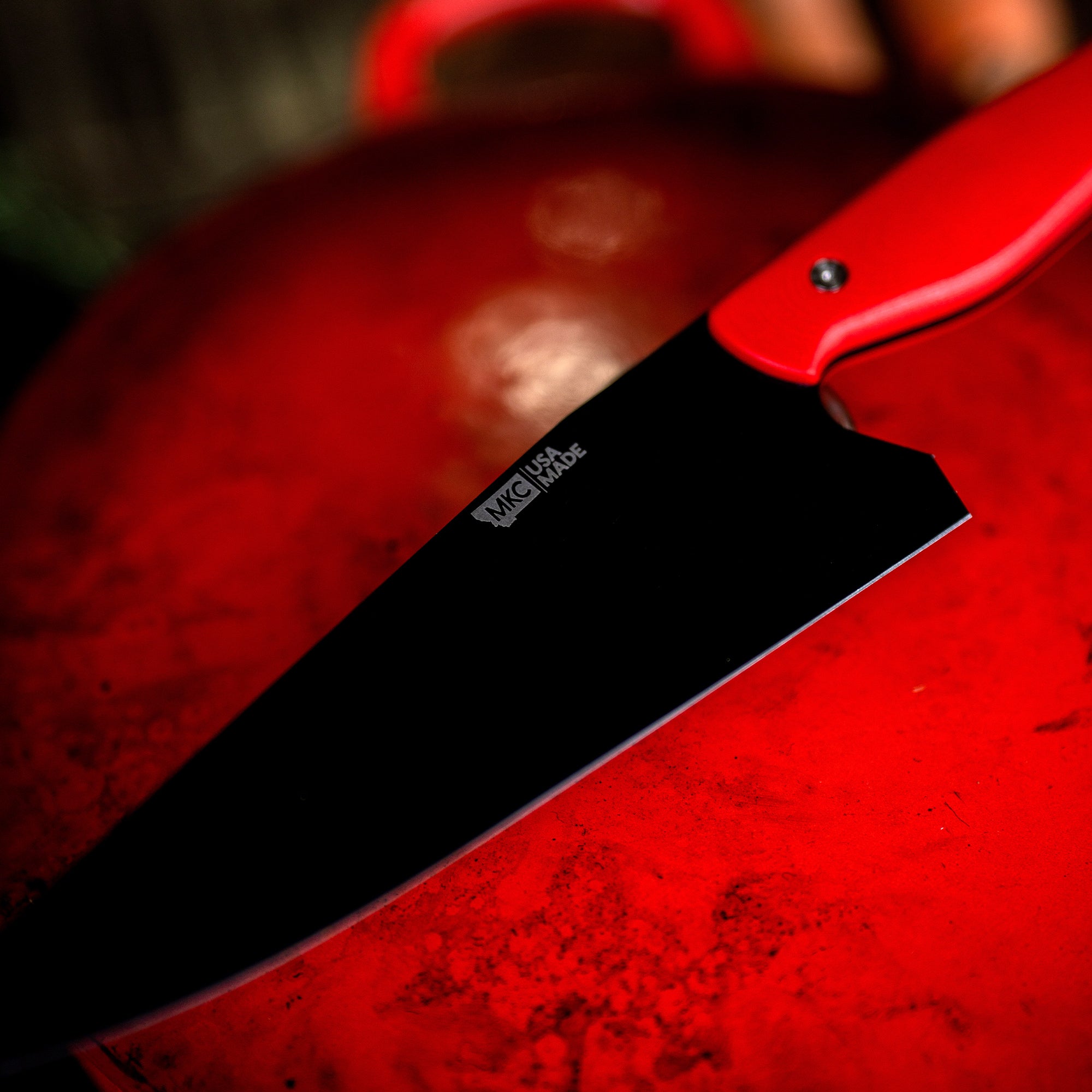 Close-up of a high-quality chef's knife with a red handle laying over a textured red surface, emphasizing the sharp, sleek black blade marked with the logo "MCUSTA."