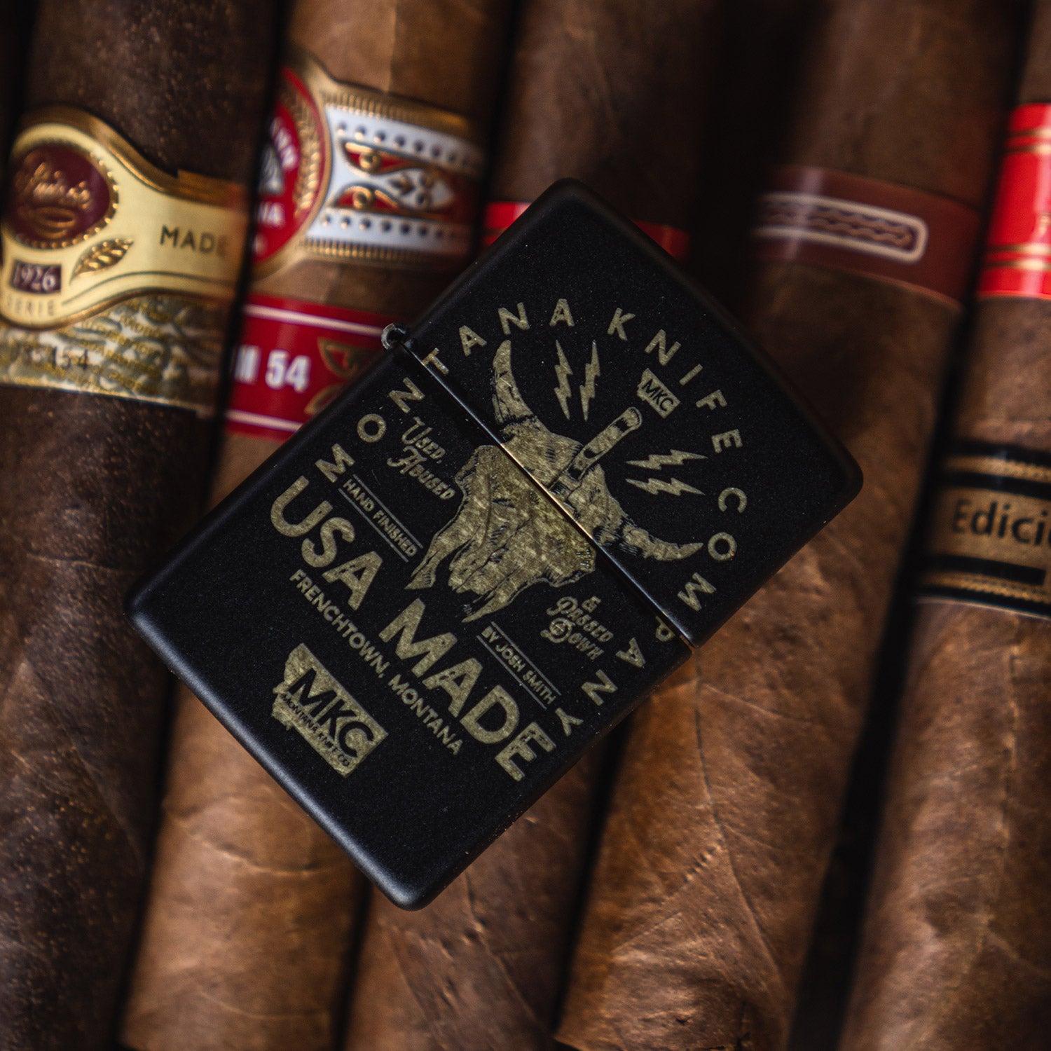 Traditional black windproof Zippo lighter featuring a detailed bison skull design and "Montana Knife Co" text, prominently displayed against a background of assorted cigars. The lighter highlights the engraving "USA Made" emphasizing its American craftsmanship.