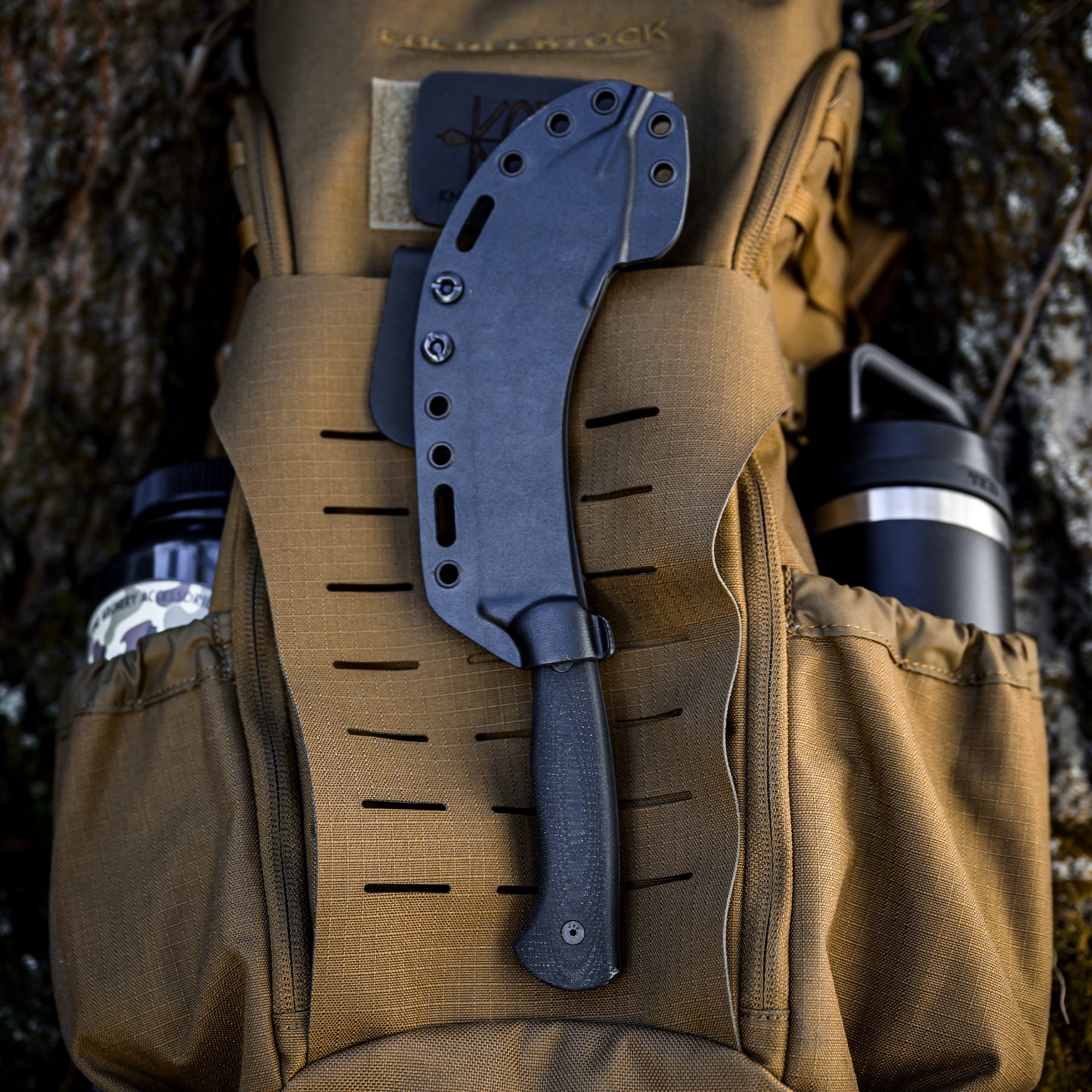 Black Beartooth Pro Skinning Blade attached to a khaki tactical backpack outdoors, featuring a secure sheath with mounting holes.