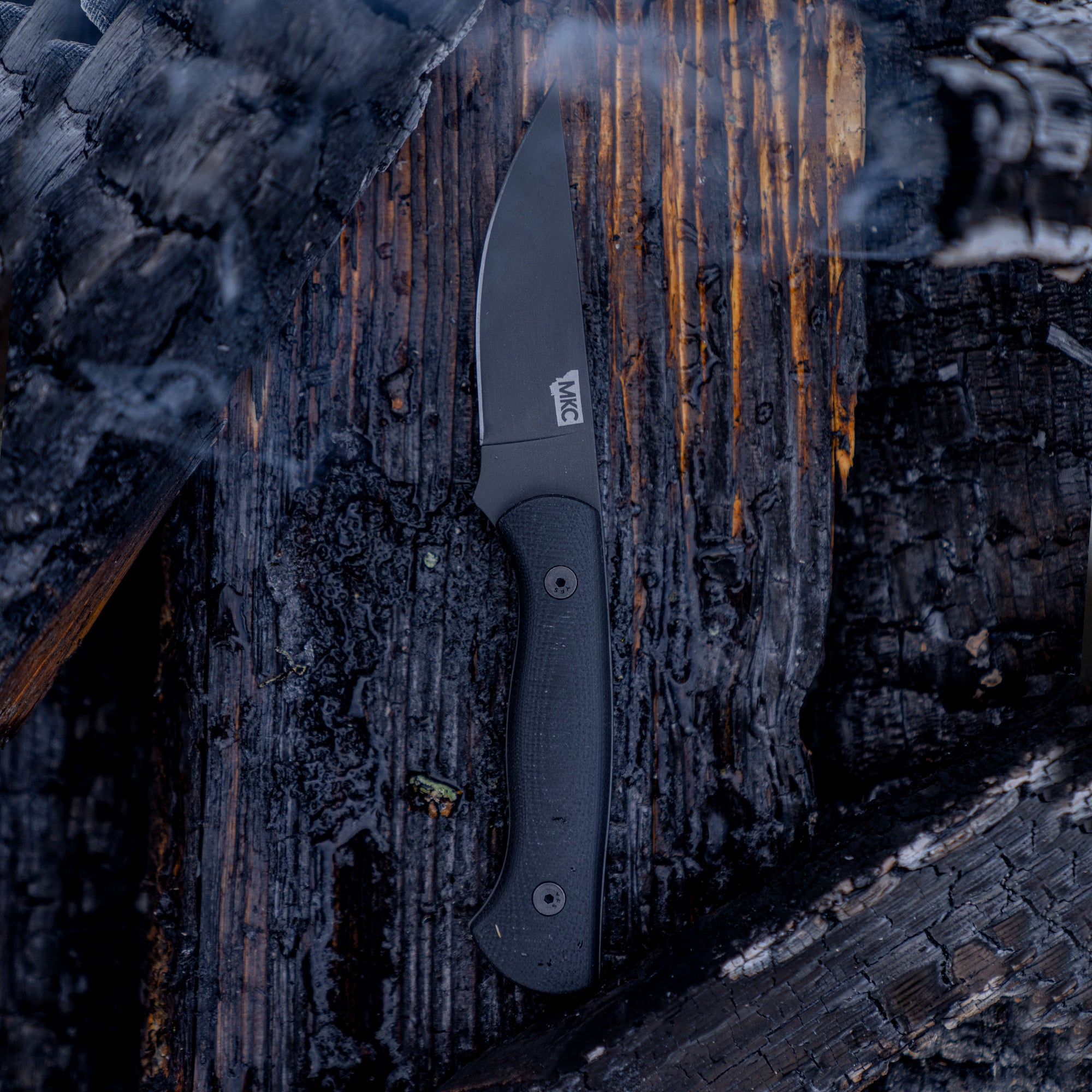 Alt text: A matte black tactical knife, the BLACKFOOT 2.0, resting on a charred log, highlighting its durable and rugged design suitable for outdoor use.