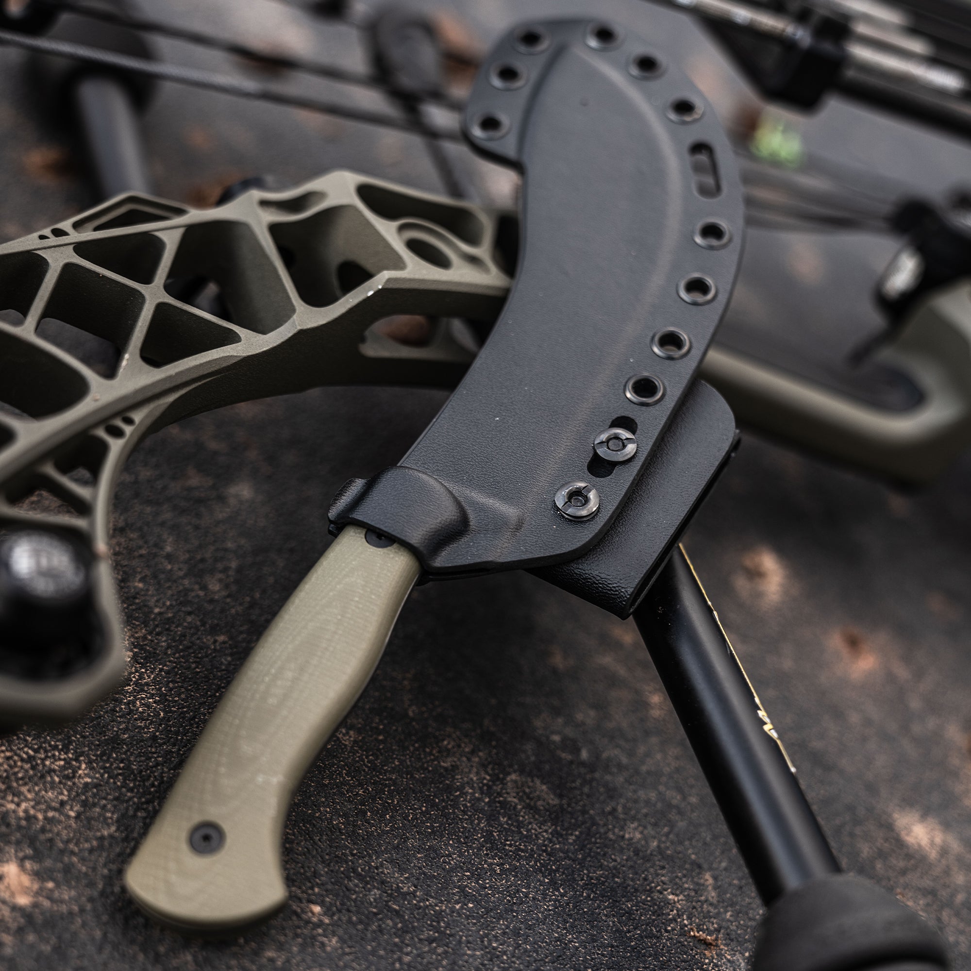 Close-up of a Beartooth Pro Skinning Blade in olive color, partially inserted into a black sheath, with a crossbow blurred in the background.