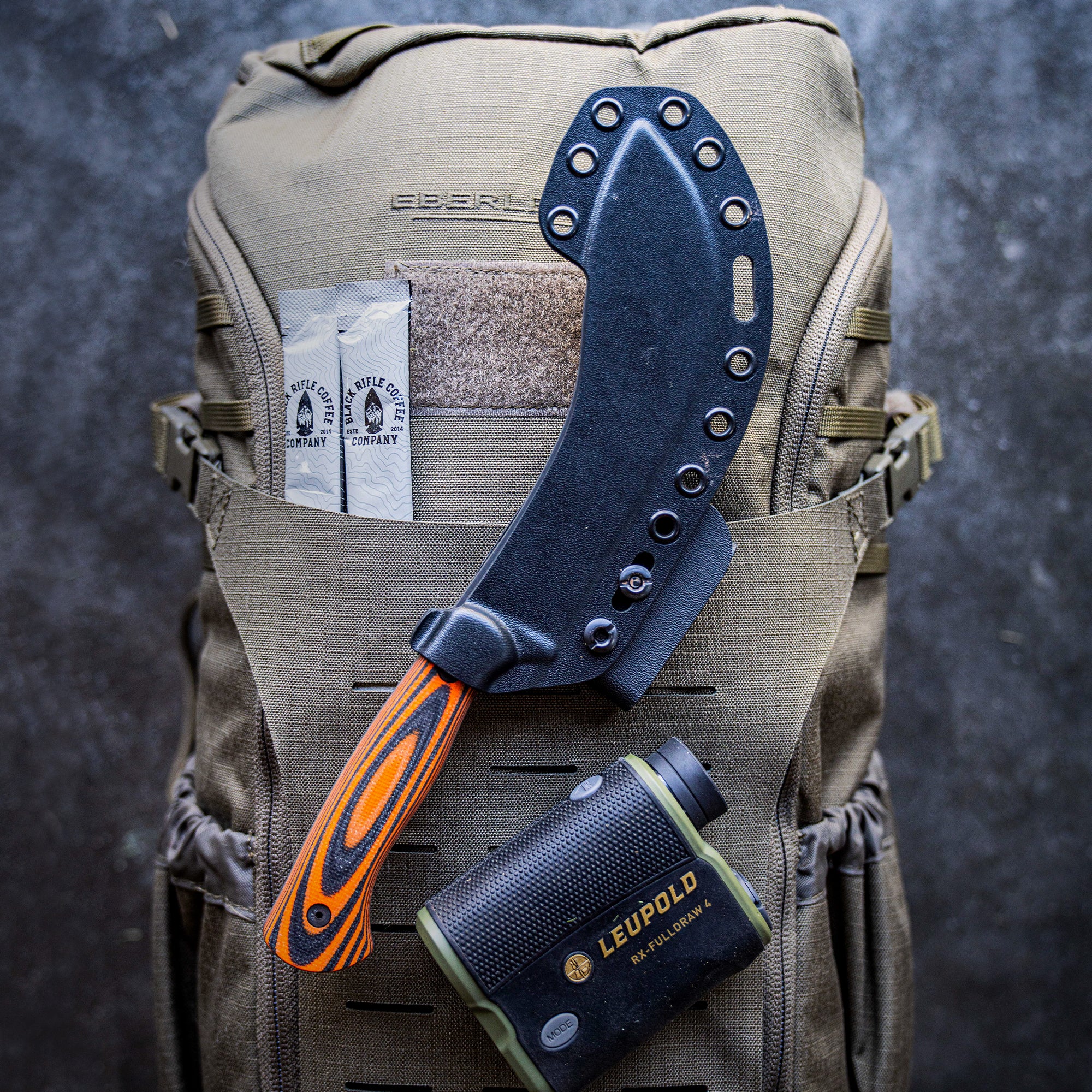Alt text: "Beartooth Pro Skinning Knife with a bright orange and black handle attached to a military green tactical backpack. The knife includes a black sheath fitted to the exterior of the backpack next to a 'Rifle Coffee Company' patch