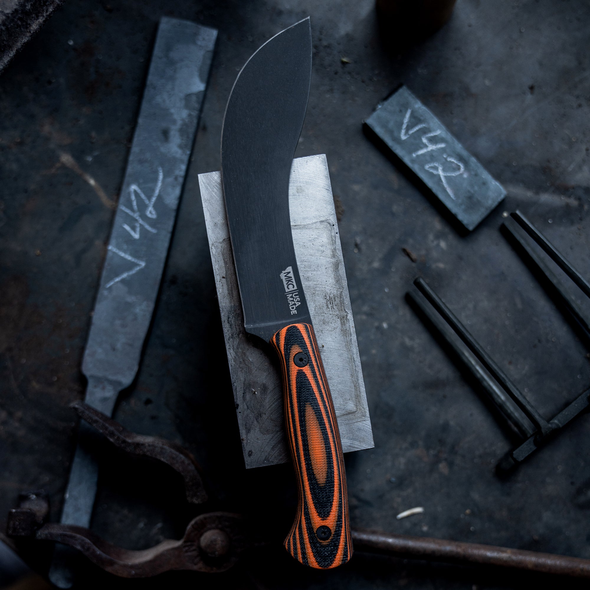 Alt text: "Beartooth professional skinning knife with a curved black blade and an orange and black handle, resting on a metallic surface in a workshop environment."