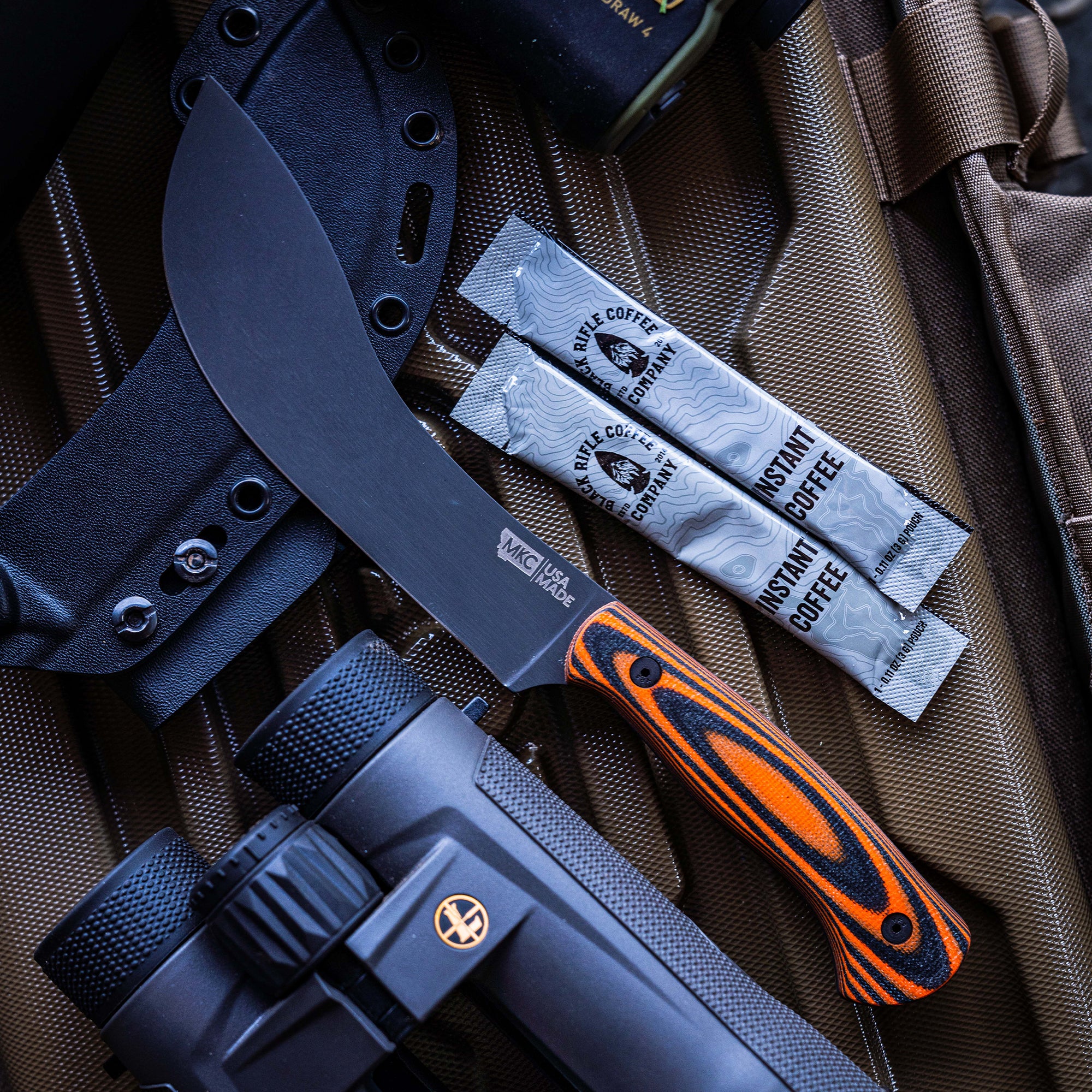 Alt text: "Close-up of a Beartooth Pro Skinning Blade with a matte black finish and orange & black handle, displayed on a brown tactical bag with a sheath, alongside a portable flashlight and packets of instant coffee."