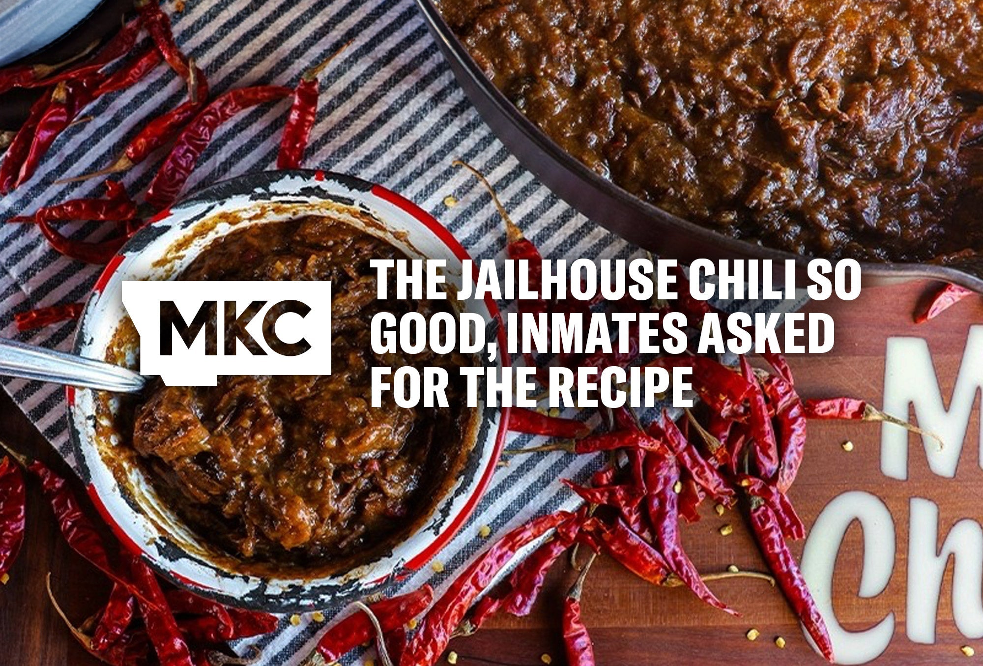Rustic enamel bowl of thick beef chili surrounded by dried red chili peppers on a striped cloth.