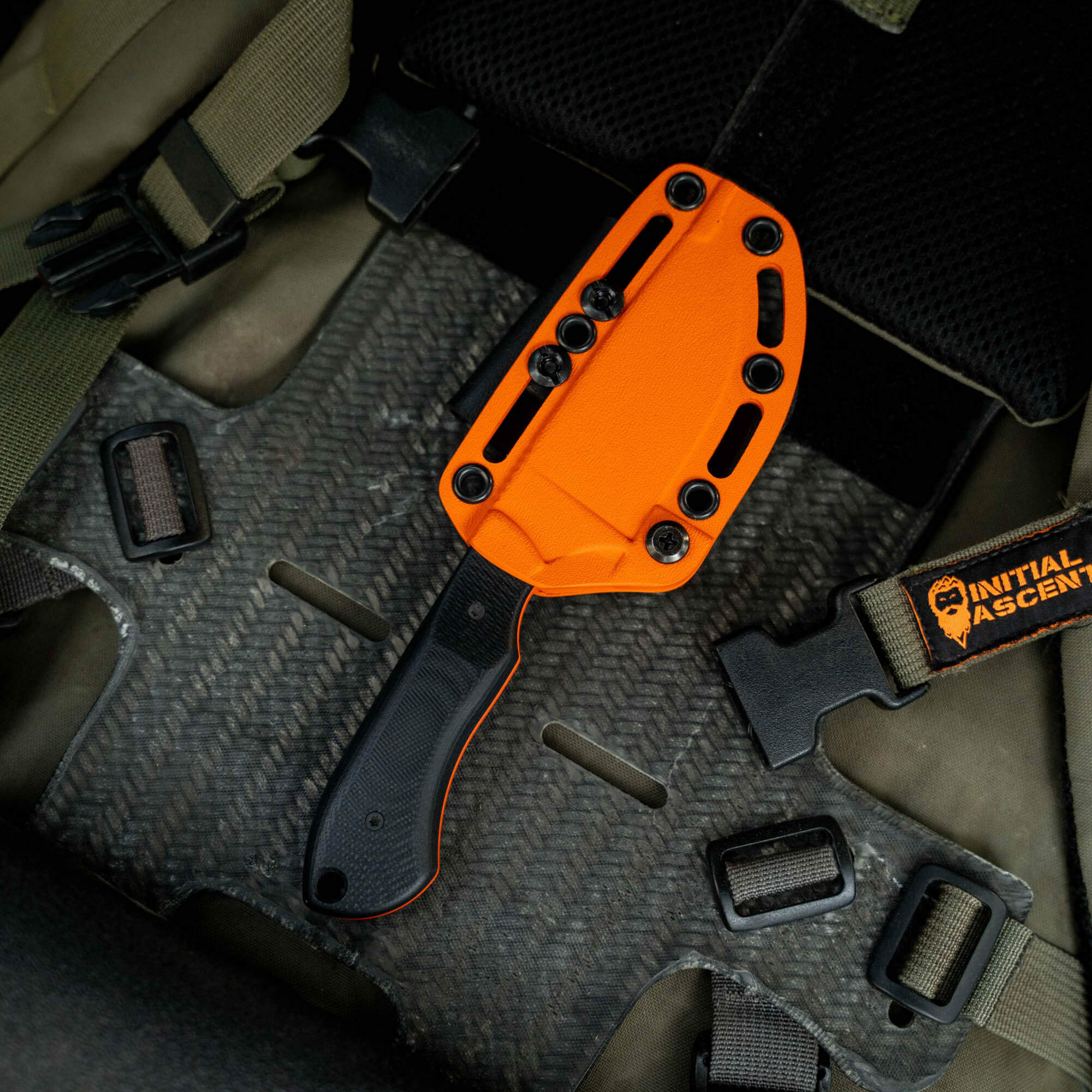 MKC Elk Knife with Blaze Orange Cerakote finish, resting in a tactical sheath on a textured surface.