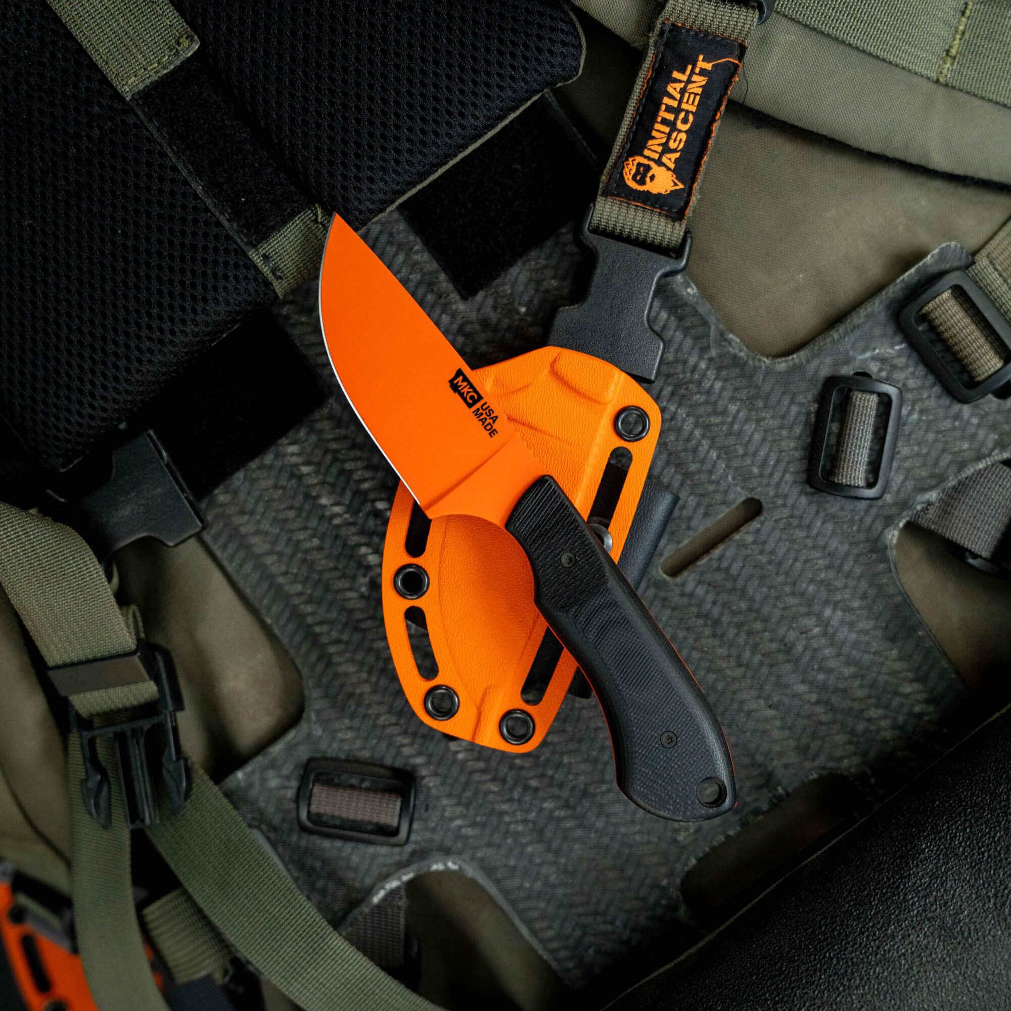 MKC Elk Knife with Blaze Orange Cerakote finish, featuring a fixed blade and textured grip, displayed on tactical gear.