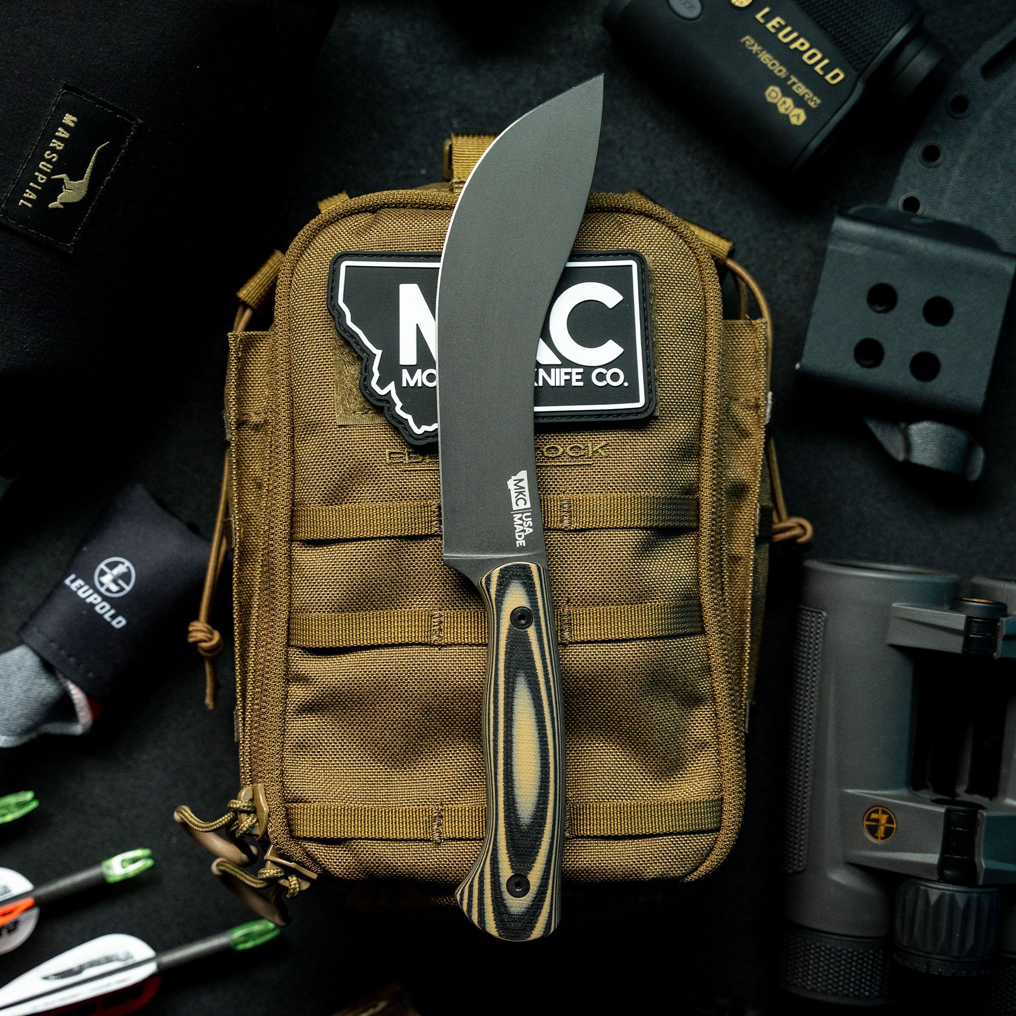 Alt text: "Beartooth Pro Skinning Blade knife with a tan and black handle, prominently displayed against a tan tactical bag with various outdoor gear items in the background, including optics and a flashlight."