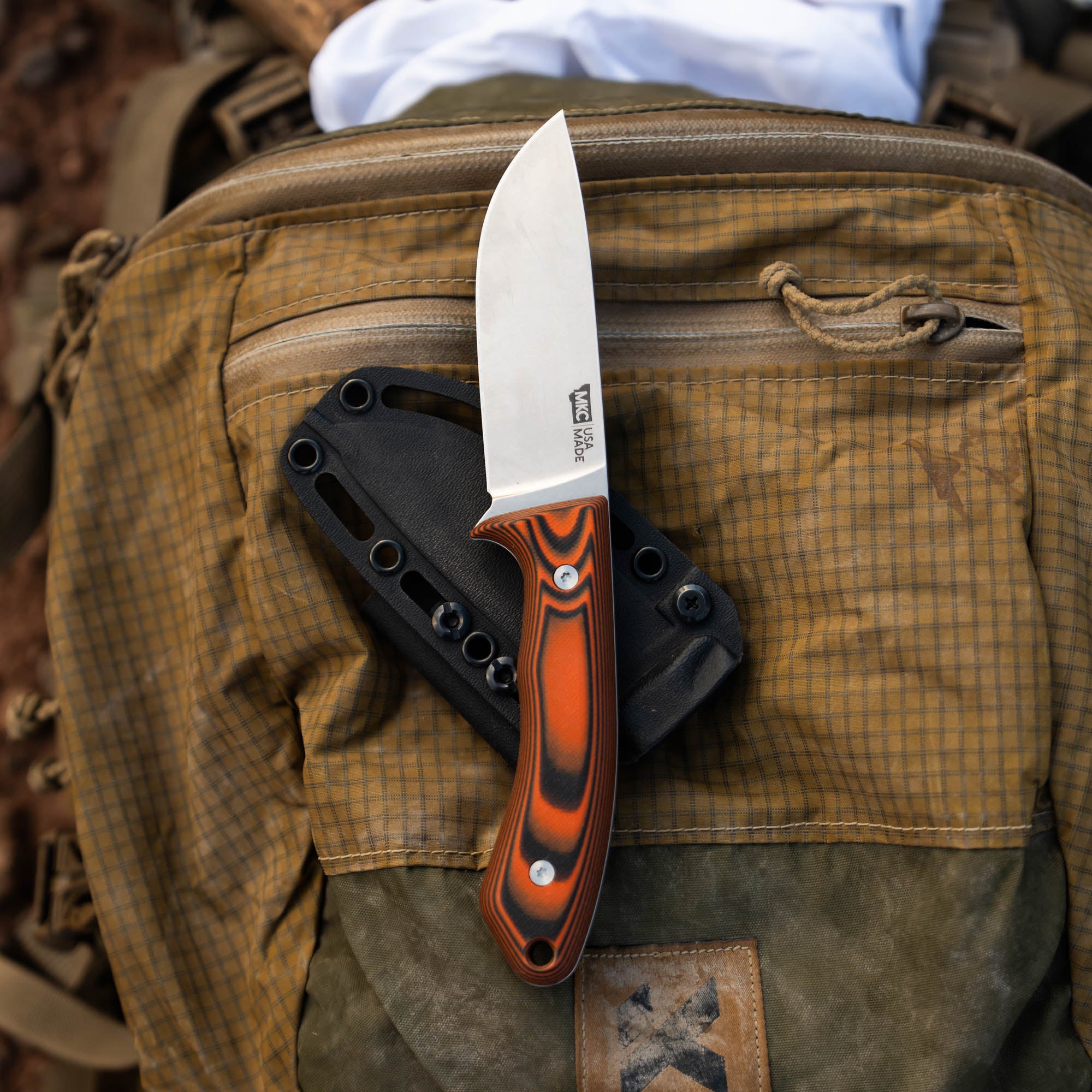 MKC Mule Deer Knife in orange and black resting on a backpack, showcasing its durable blade and ergonomic handle.