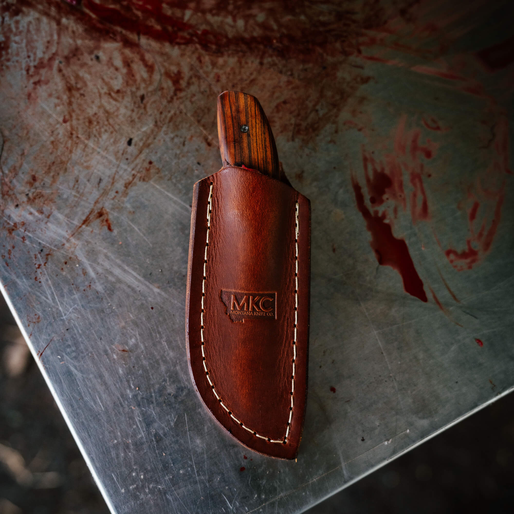 Montana Knife Company's Keep Hammering Traditions Packout Skinner in a leather sheath, showcasing craftsmanship and durability.