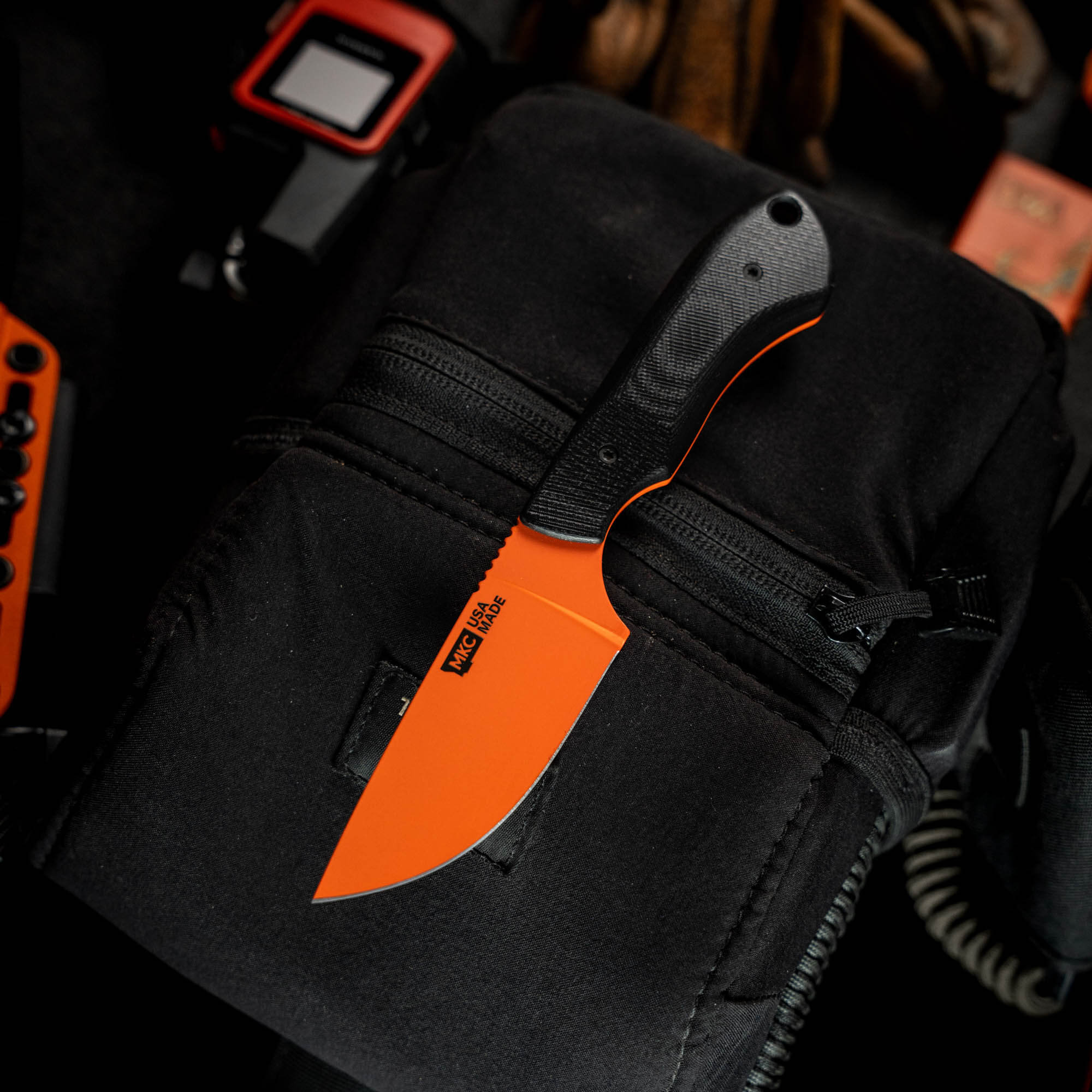 MKC Elk Knife in Blaze Orange Cerakote with a black handle, resting on a tactical pouch. Ideal for hunting and outdoor use.
