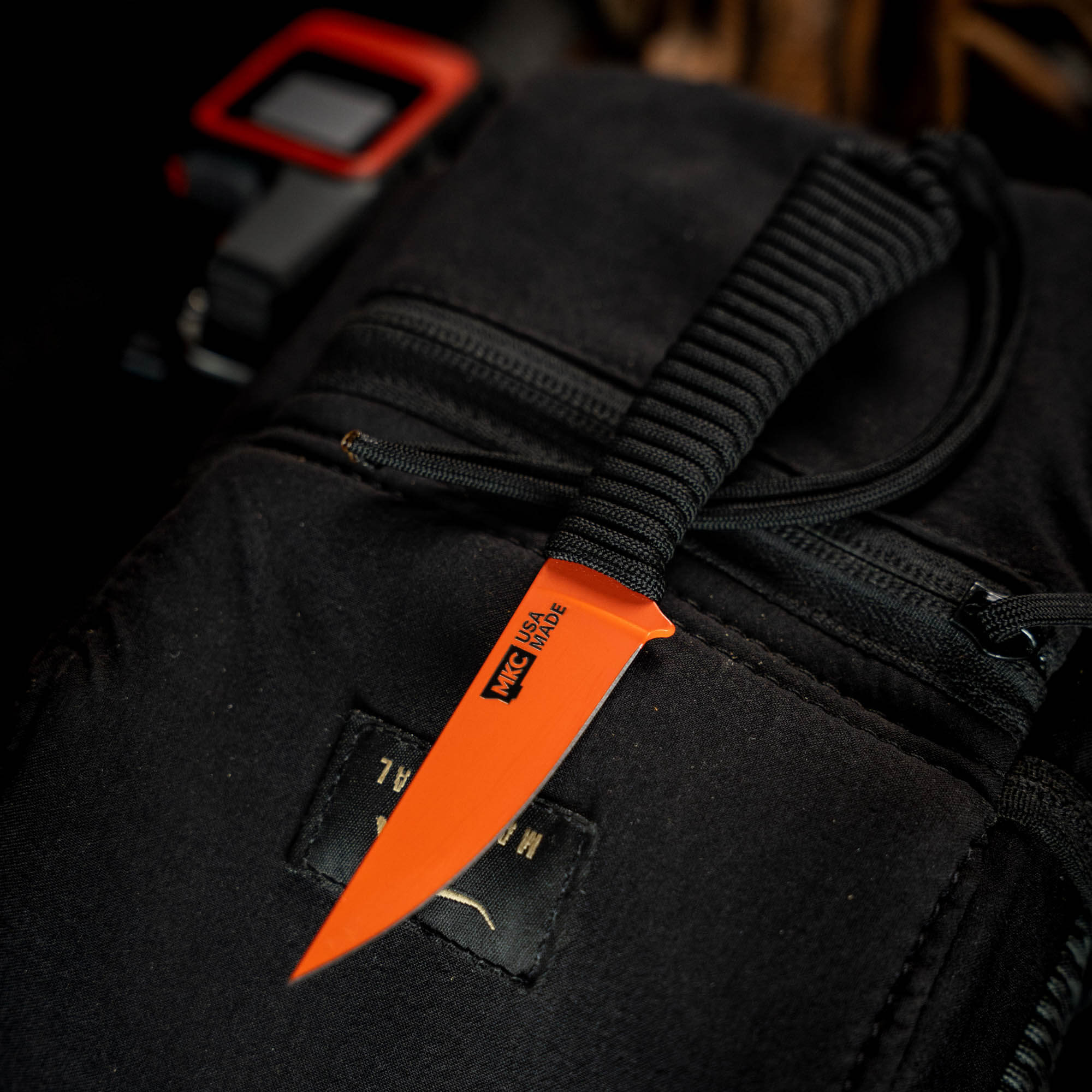 Mini Speedgoat 2.0 knife in Blaze Orange Cerakote, featuring a durable blade and paracord handle, resting on a black backpack.