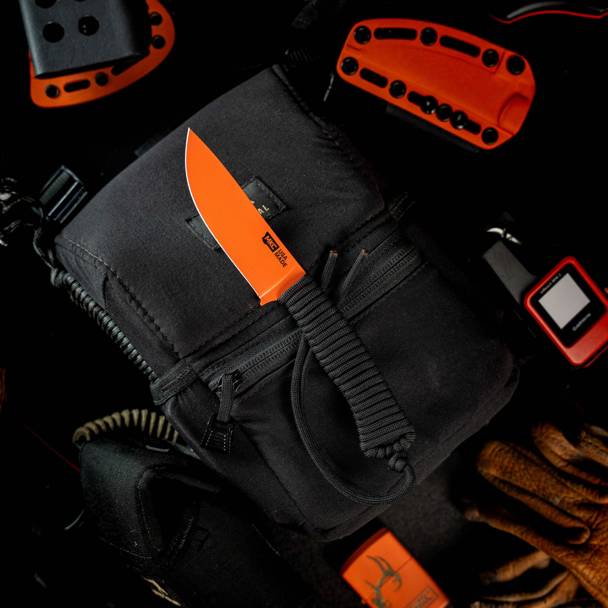 Blaze orange Cerakote Stoned Goat 2.0 knife on a black tactical bag, surrounded by outdoor gear and tools.