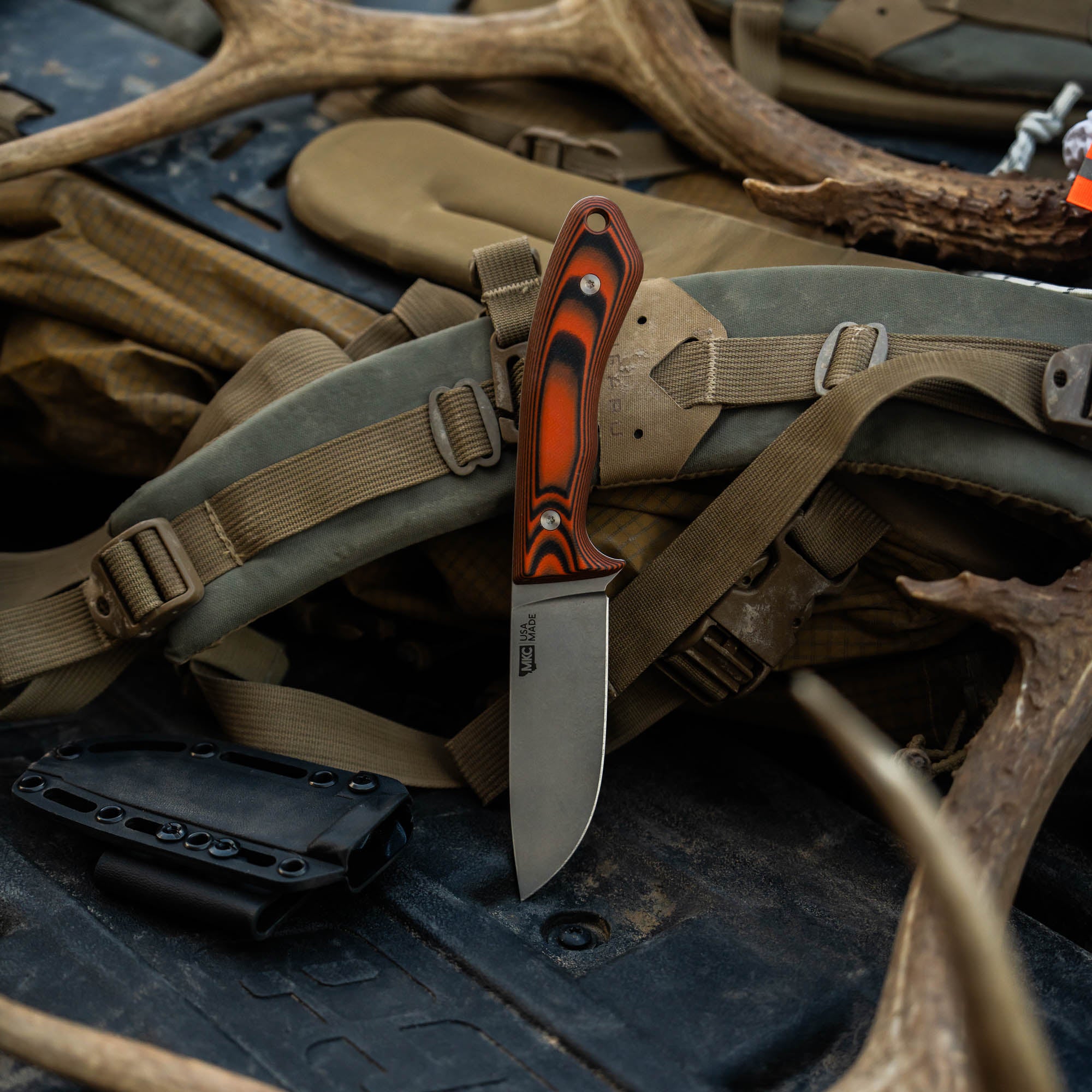 MKC Mule Deer Knife in orange and black resting on gear with antlers, showcasing its rugged design for outdoor use.
