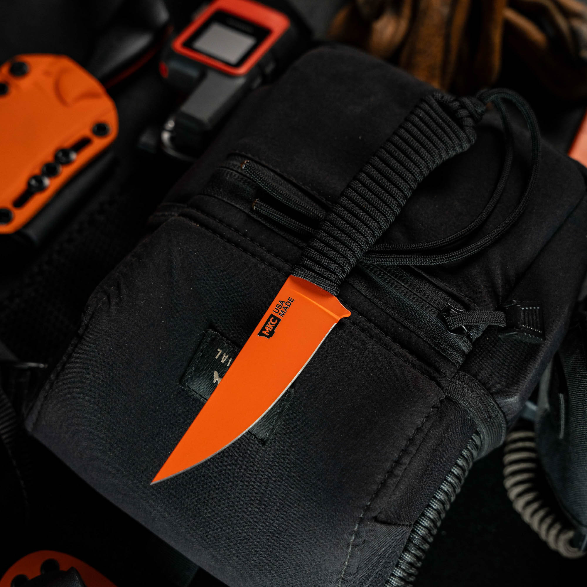 SPEEDGOAT 2.0 knife in blaze orange Cerakote, resting on a black tactical pouch with outdoor gear.