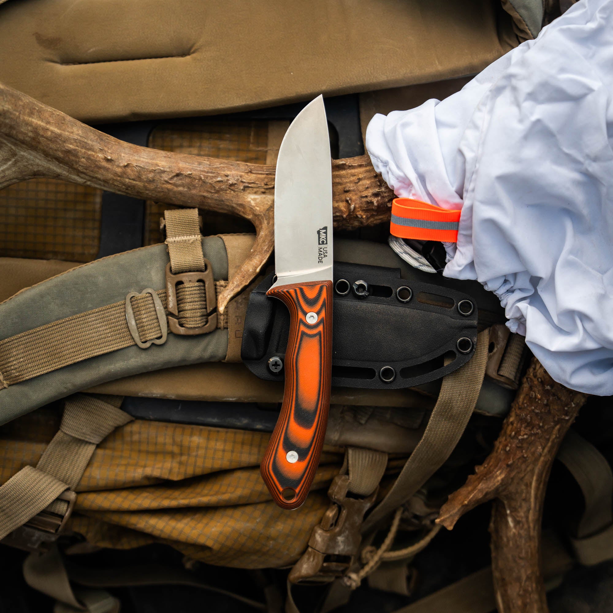 MKC Mule Deer Knife in orange and black resting on a tactical backpack, showcasing its fixed blade and sheath.