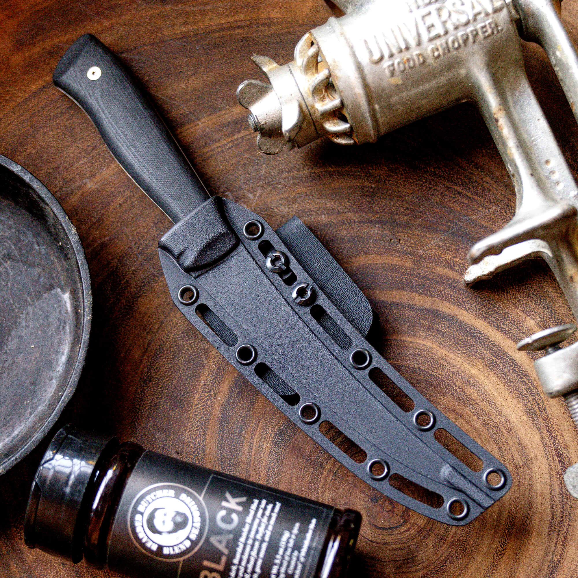 Alt text: "Bearded Butchers boning butcher knife in a black sheath, displayed on a wooden surface alongside a vintage Universal food chopper and a bottle of Black Reserve seasoning."